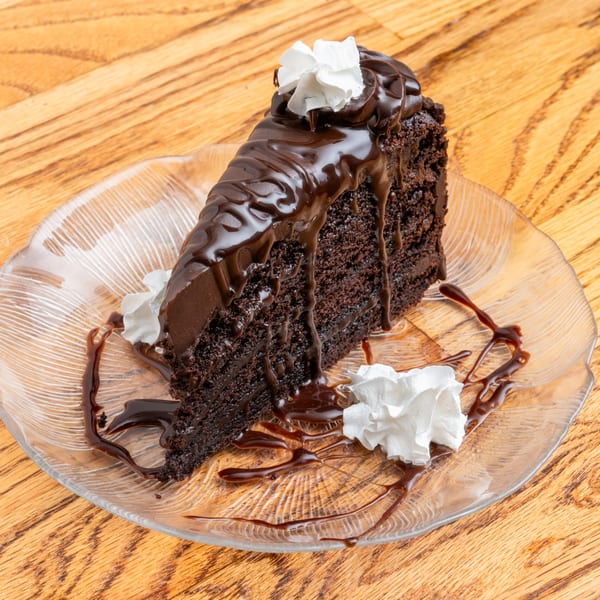 A slice of rich chocolate cake with chocolate ganache and whipped cream on a clear plate.